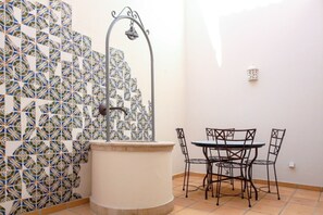 Un mur recouvert de carreaux formant un motif géométrique, une fontaine à eau en métal, une table ronde avec des chaises, et un mur blanc avec un interrupteur.