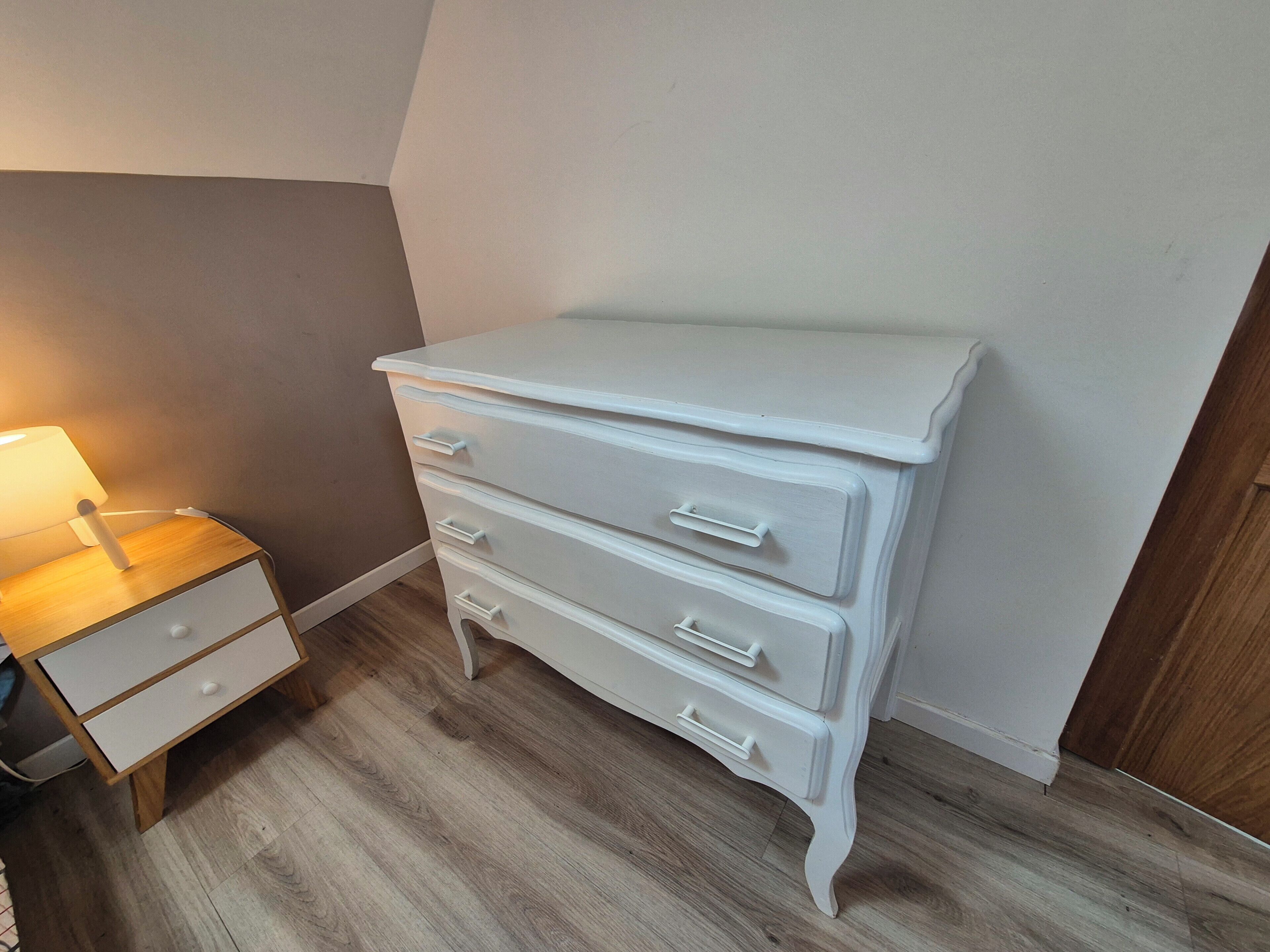 Un aparador blanco con cajones, una mesita de noche con lámpara y suelo de madera.