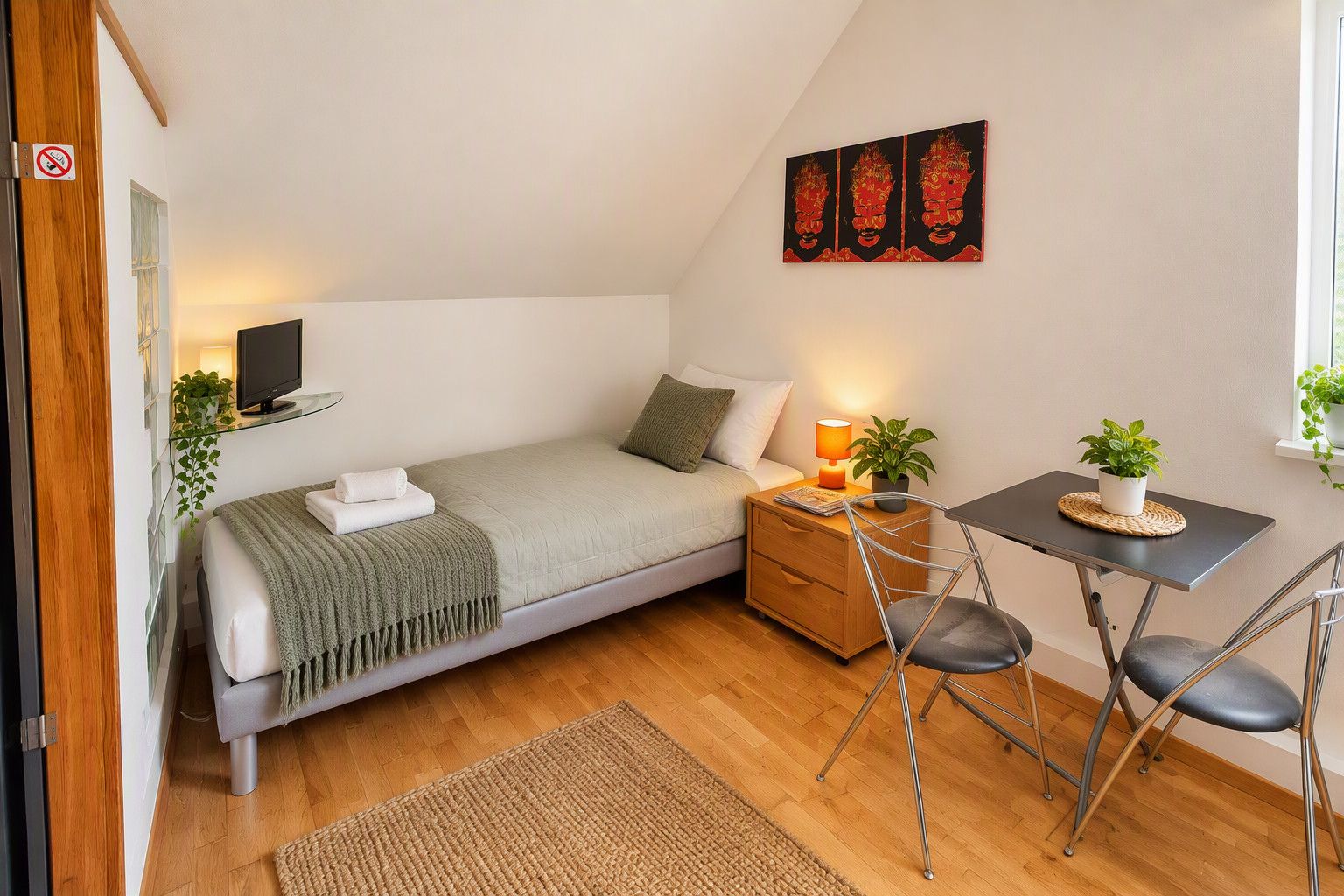 Une petite chambre à coucher accueillante, avec un lit, une table de chevet, une lampe, une chaise et un téléviseur fixé au mur.