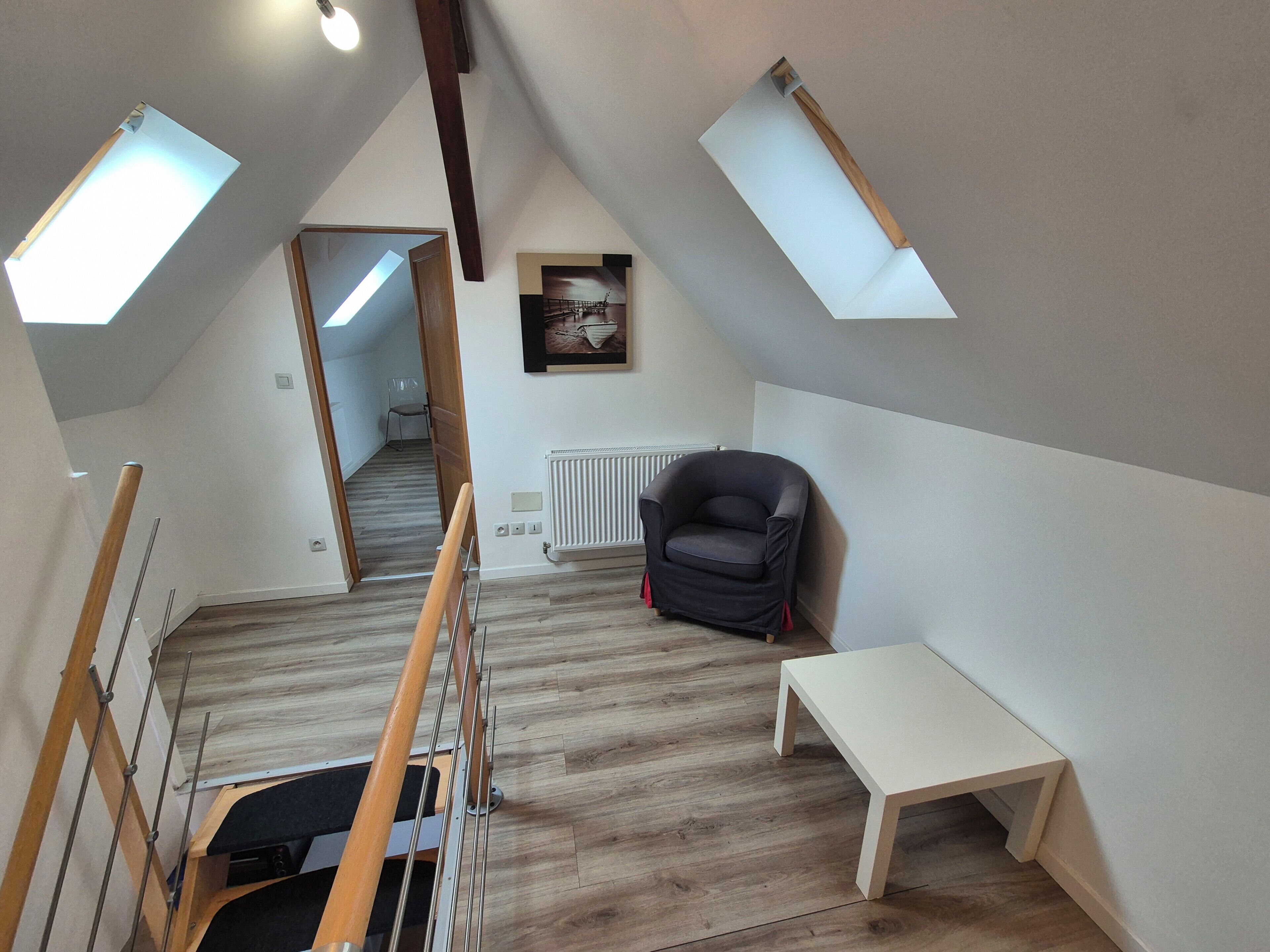Ein Schlafzimmer im Loft-Stil mit schrägen Wänden, Dachfenstern, einer Heizung, einem schwarzen Stuhl und einem kleinen Tisch.