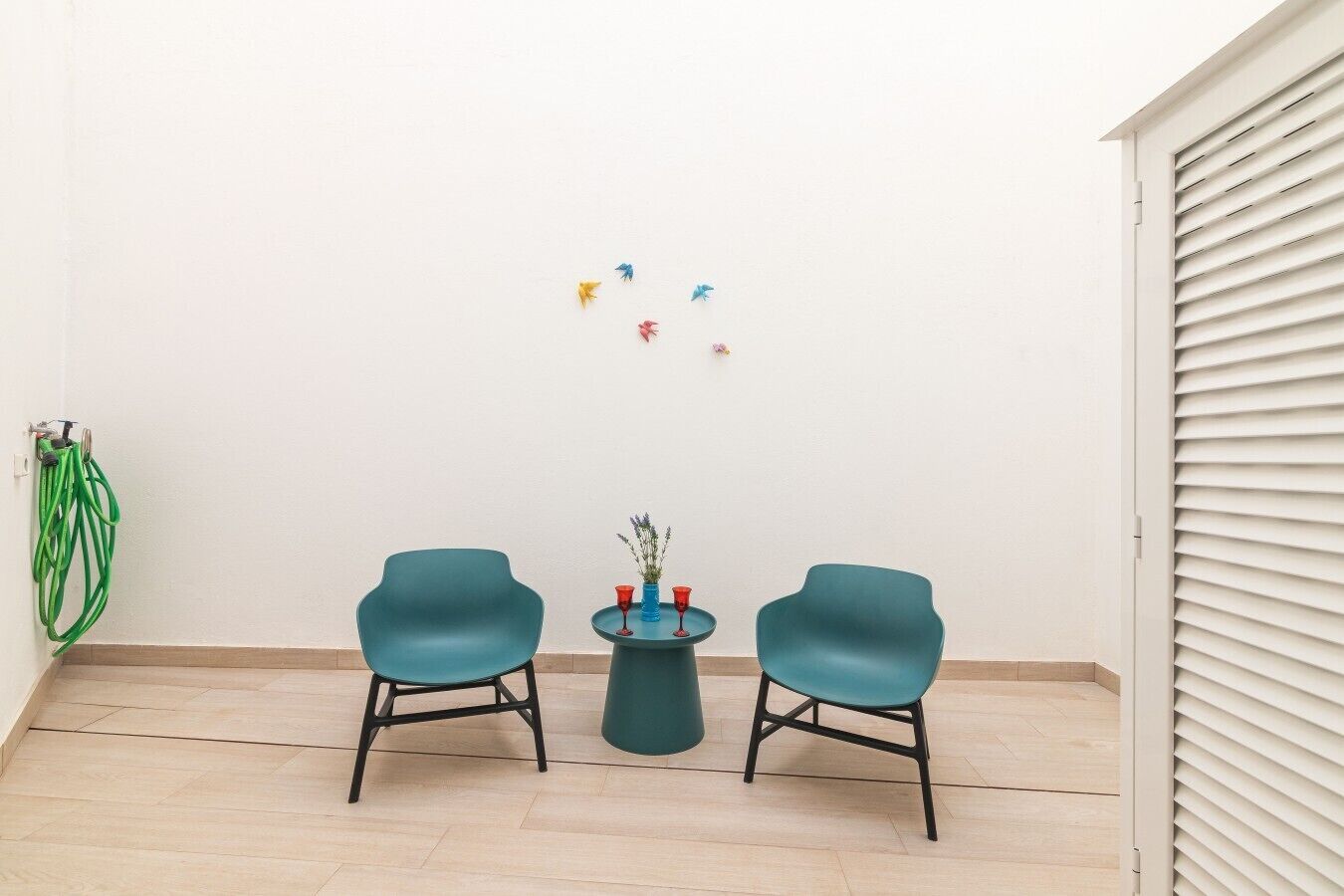Ein Zimmer mit zwei Türkis-Stühlen, einem kleinen runden Tisch mit einer Vase und einer roten Tasse, sowie einer weißen Wand mit einem grünen Schlauch links.