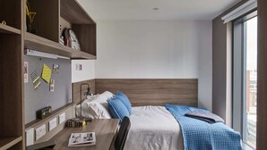 A bedroom with a bed, a desk with a chair, a window, and a wall with a clock and photos. - Student Only Howard Gardens (Cardiff)