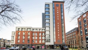 A multi-story building with a prominent 'HOWARD GARDENS' sign, featuring a mix of brick and glass facades, and several ground-level retail spaces. - Student Only Howard Gardens (Cardiff)