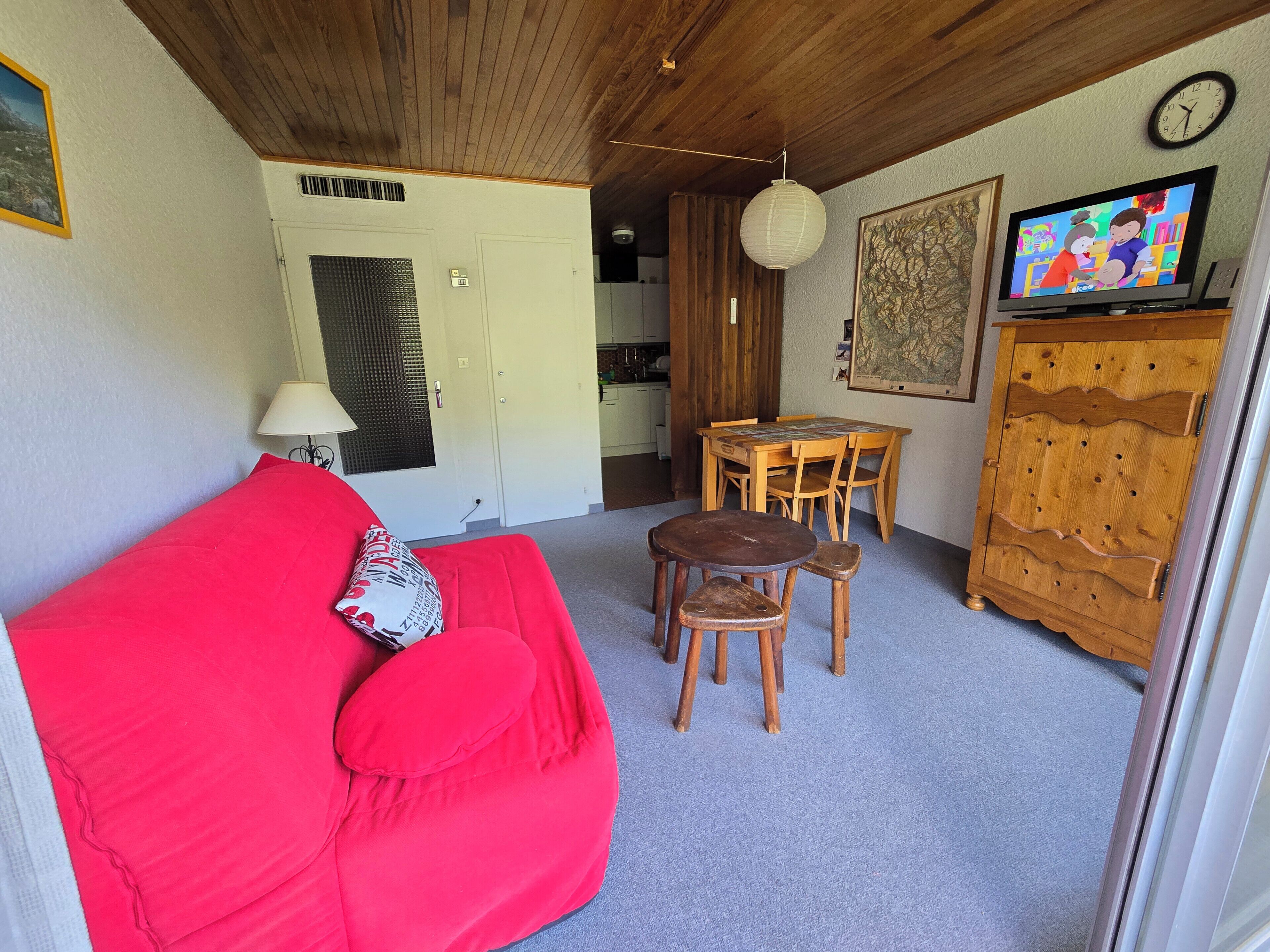 A cozy living area with a red sofa, a wooden dining table with chairs, a television, and a wooden cabinet.