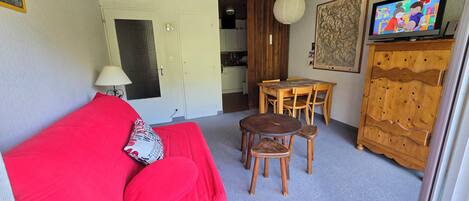 A cozy living area with a red sofa, a wooden dining table with chairs, a television, and a wooden cabinet.