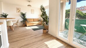 Ein modernes Wohnzimmer mit Holzboden, einem beigen Sofa, einem Esstisch mit Stühlen und einer großen Glasschiebetür, die zu einem Außenbereich führt.