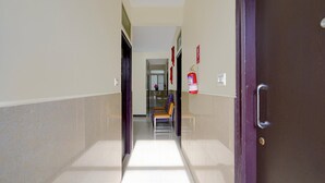 A corridor with a fire extinguisher mounted on the wall, a door on the right, and a bench in the distance. - Classic Red Cherry Wagtail (Bengaluru)