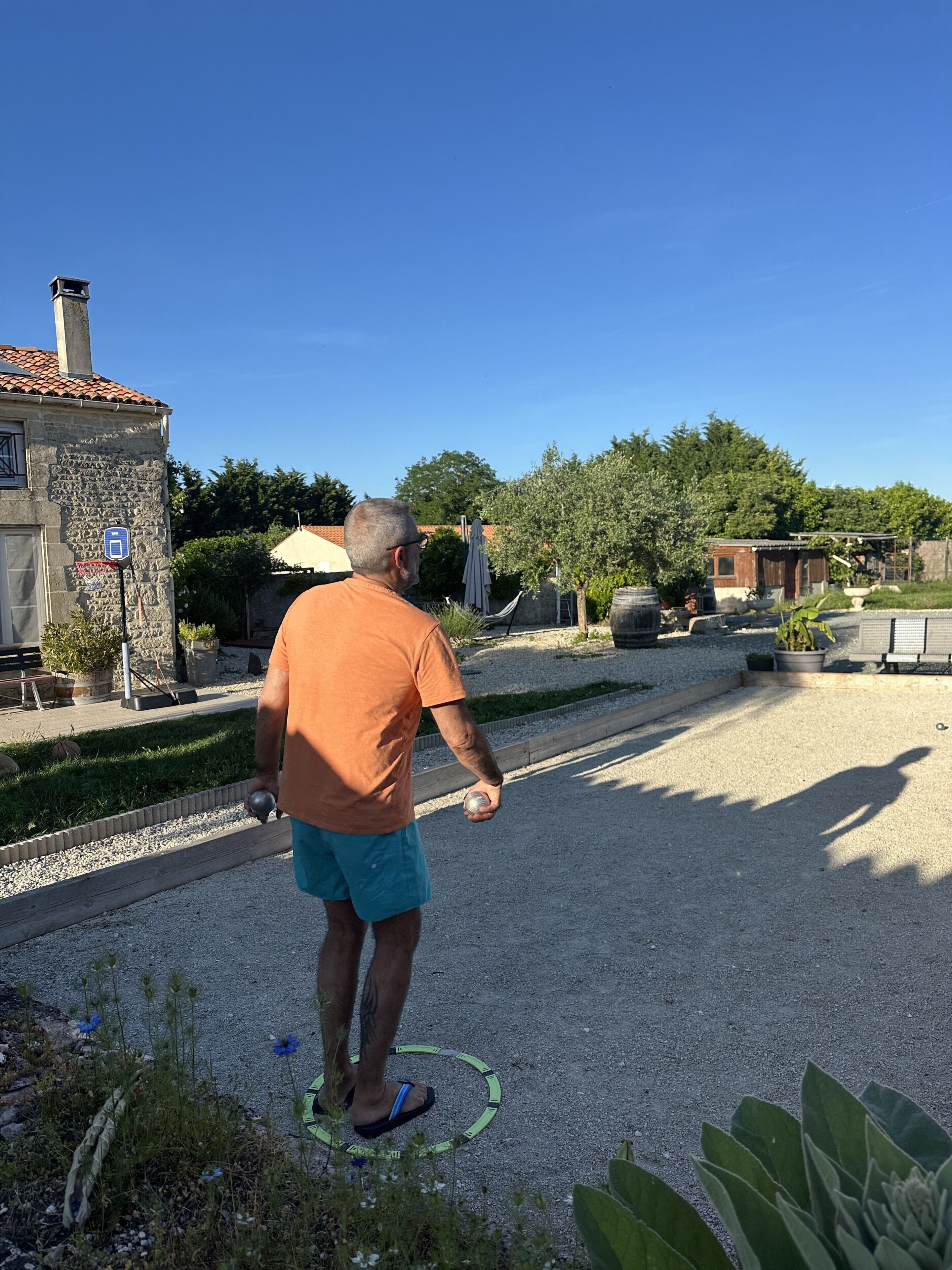 A man walking on a gravel path with a hula hoop, surrounded by greenery and a building with a stone facade.