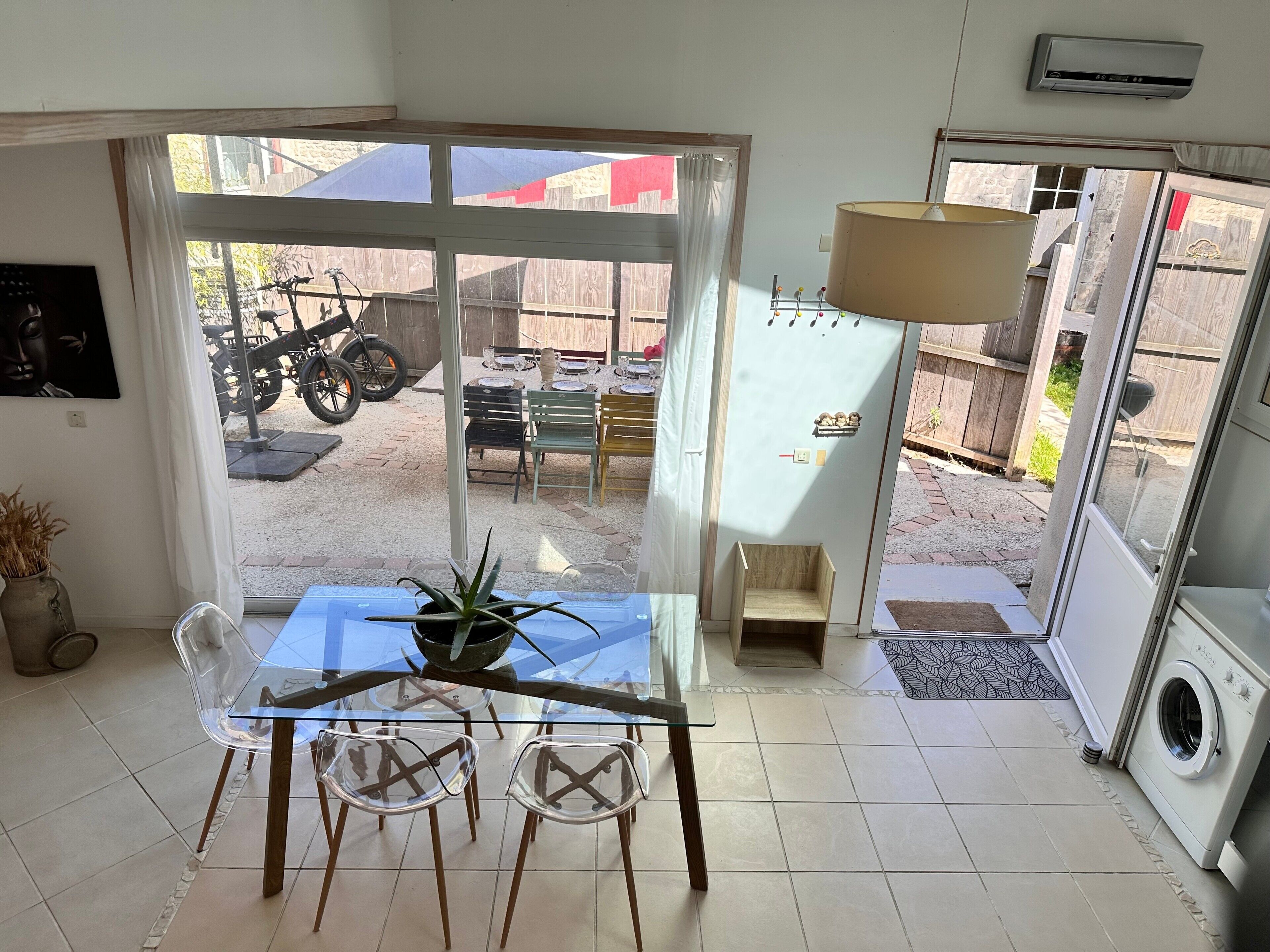 A room with a glass table, chairs, a potted plant, a washing machine, and a view of a patio with a motorcycle and outdoor furniture.