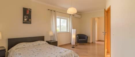 Un dormitorio con cama, mesitas de noche, una silla, una ventana con cortinas y un equipo de aire acondicionado.