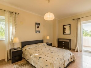 Ein Schlafzimmer mit einem Bett, Nachttischen, einer Hängeleuchte, einer Kommode und einem Fenster mit Vorhängen.