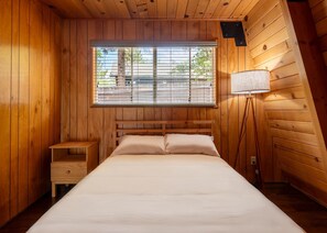 Ein Zimmer mit Holzboden, einem Bett, einem Nachttisch, einer Lampe und einem Fenster mit Jalousien.