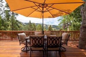 Eine Holzterrasse mit Tisch und Stühlen unter einem großen Sonnenschirm, mit Blick auf eine gebirgige Landschaft.