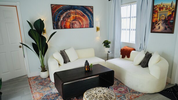 A living room with a white sofa, a dark coffee table, a patterned ottoman, a large potted plant, and two wall-mounted light fixtures. - Private Pool Paradise Near Hard Rock & Beach (Hollywood)