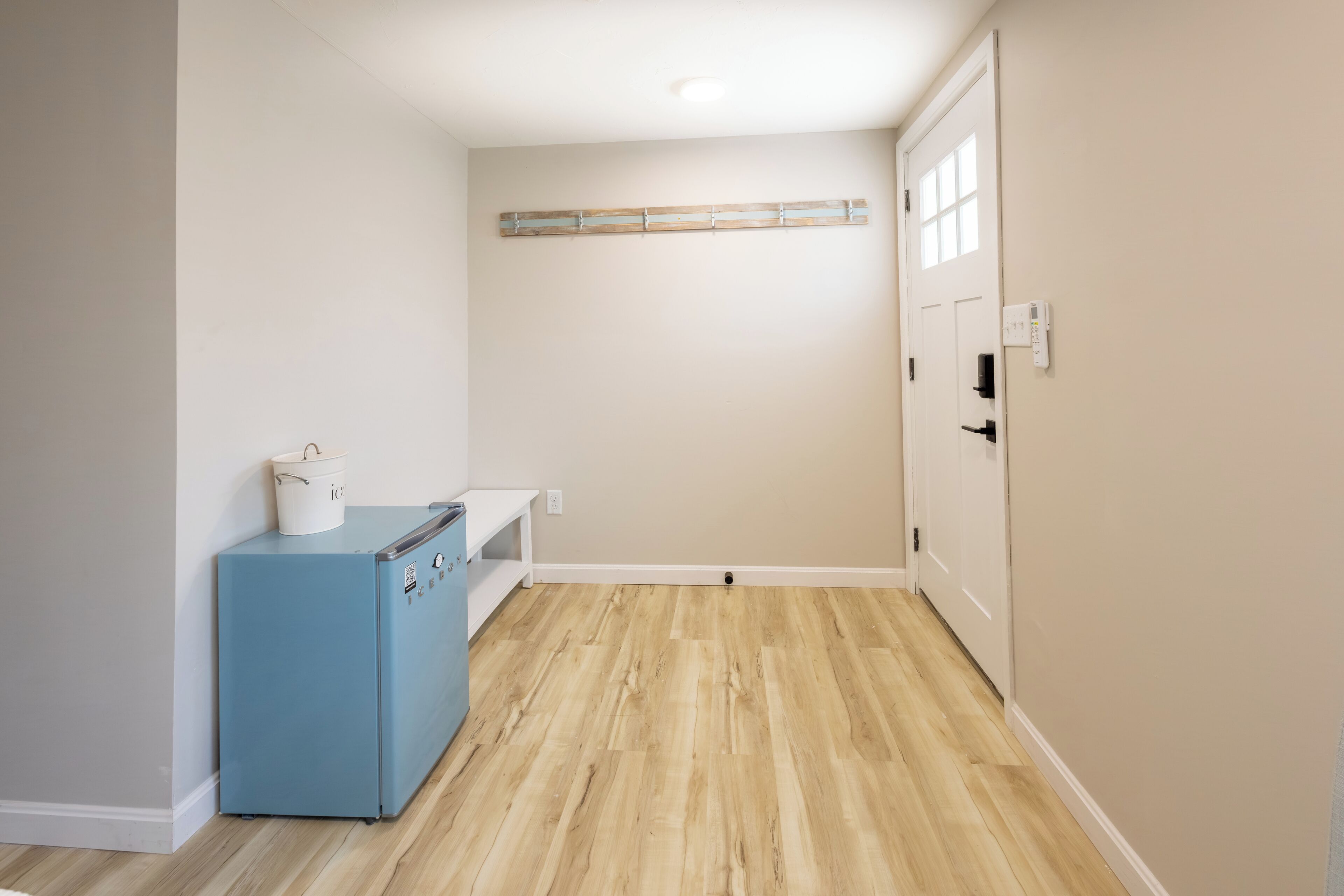 A room with a wooden floor, a blue appliance, a white bench, and a door with a light switch.