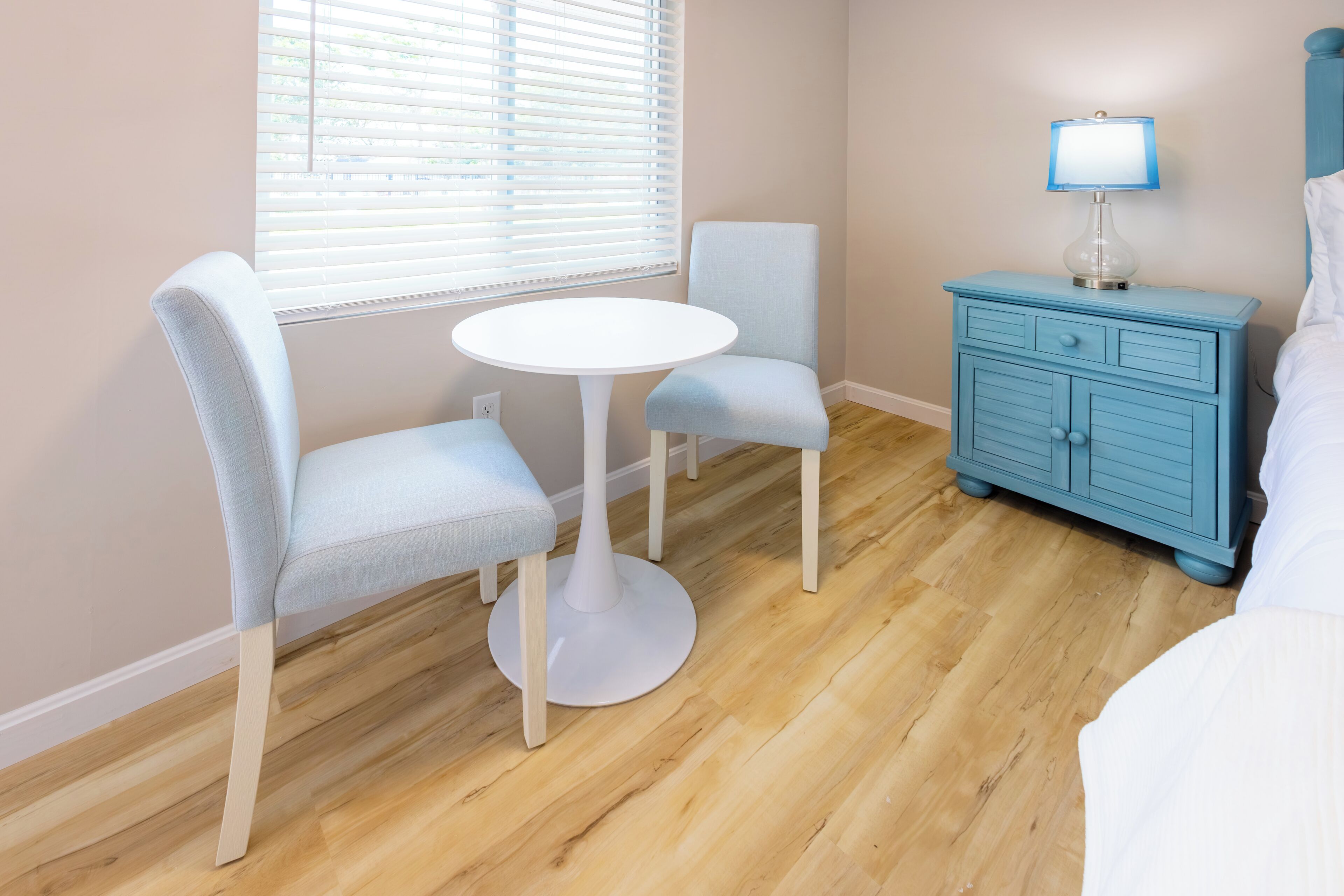 A room with a white round table, two light blue chairs, a blue nightstand with a lamp, and a bed with white bedding.