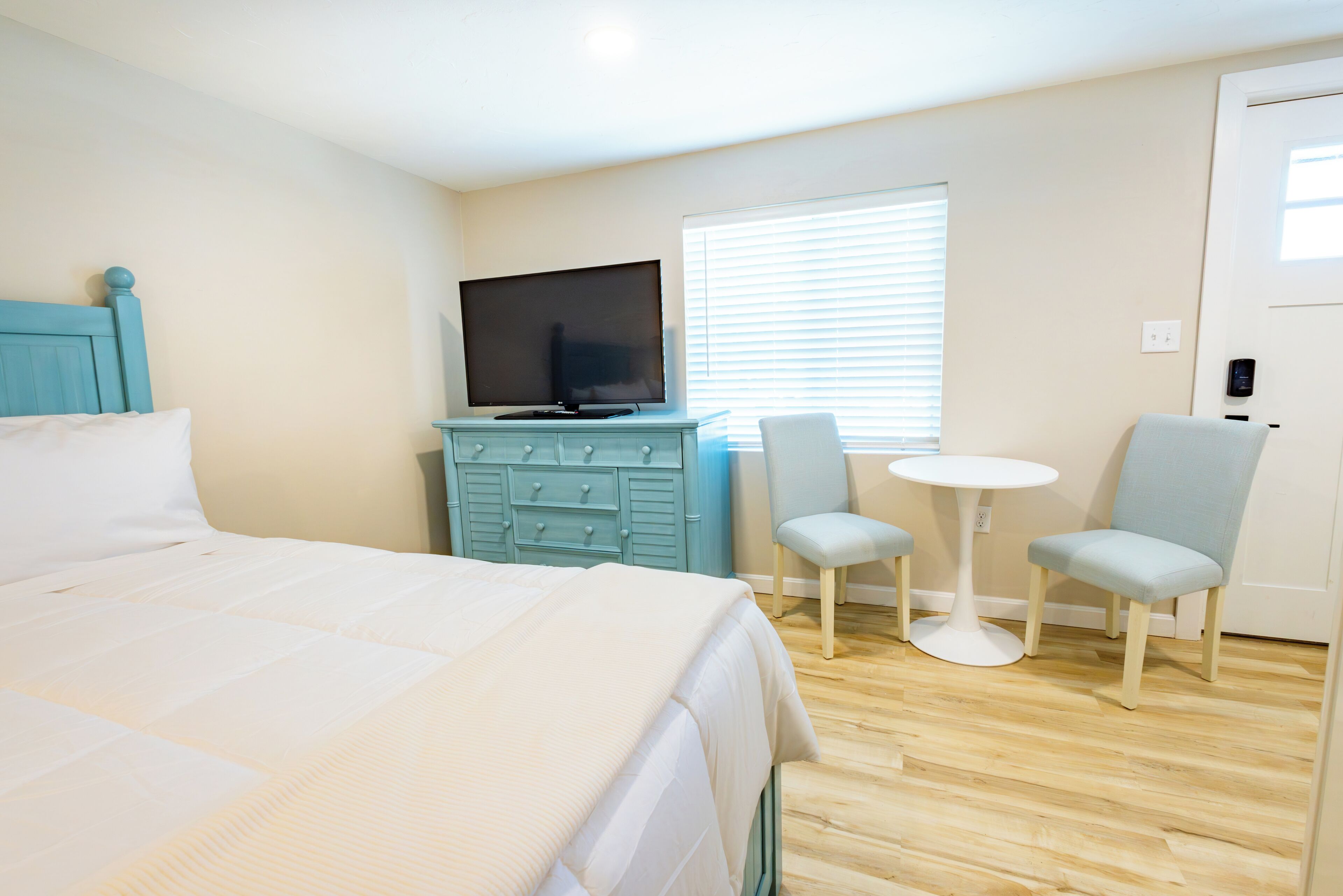 A bedroom with a bed, a TV on a dresser, two chairs, a small table, and a window with blinds.