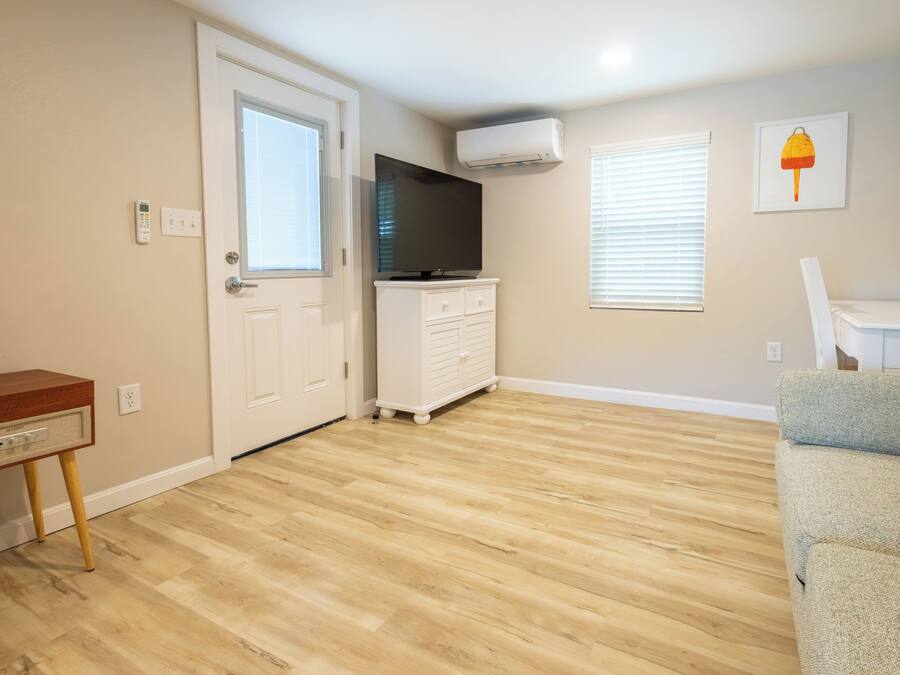 Una habitación con piso de madera, una puerta blanca, un tocador blanco, un televisor de pantalla plana y una ventana con persianas.