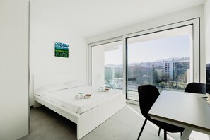 Ein modernes Schlafzimmer mit Bett, Esstisch und großem Fenster mit Blick auf die Stadt.
