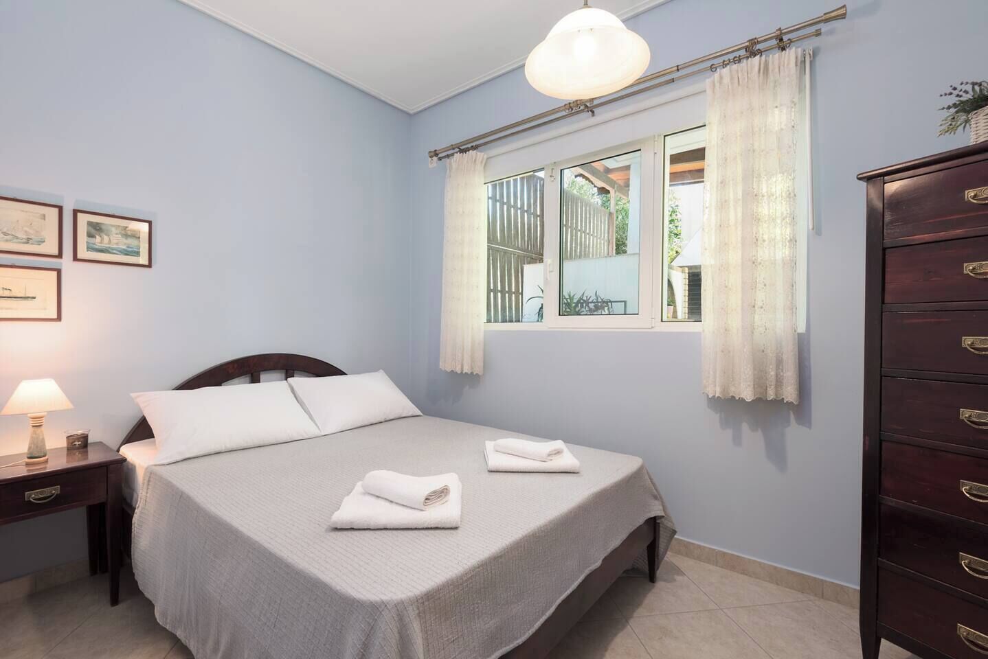 A bedroom with a bed, a nightstand, a dresser, a window with curtains, and framed pictures on the wall.