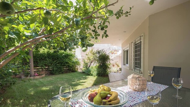A glass table with a fruit bowl, two wine glasses, and a woven basket in a sunny outdoor area with a view of a building. - Lagonisi Garden Stay, 10min Walk to Beach (Kalyvia Thorikou)