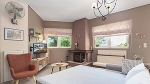A bedroom with a bed, a chair, a small table, a fireplace, and a window with a view of greenery. - Lagonisi Garden Stay, 10min Walk to Beach (Kalyvia Thorikou)