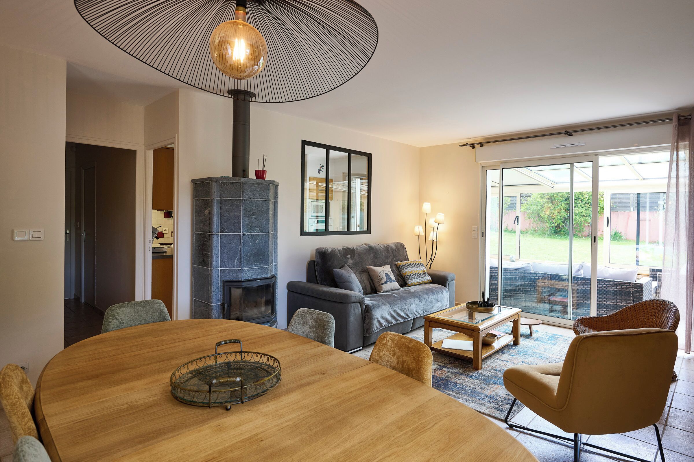 A modern living room with a round wooden dining table, a fireplace, a sofa, and a large window with a view of the outdoors.