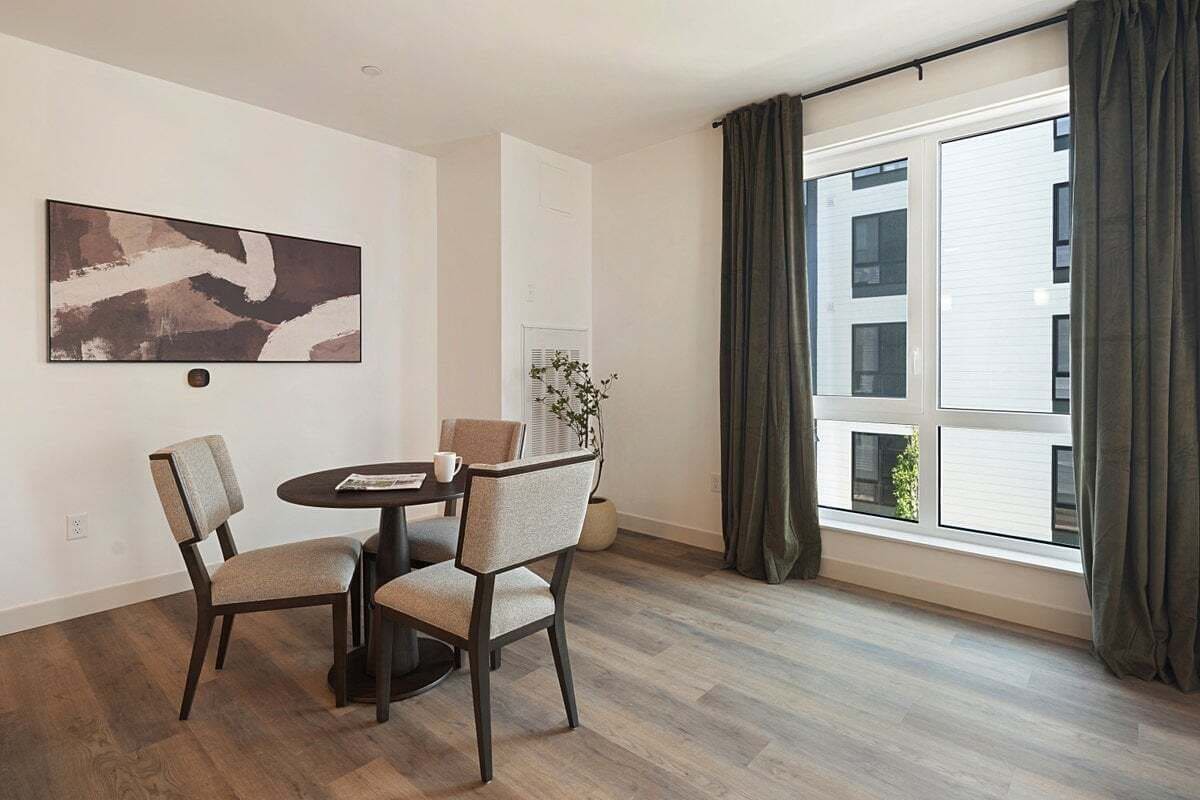 A modern dining area with a round table, two chairs, a wall-mounted abstract painting, and a large window with curtains.