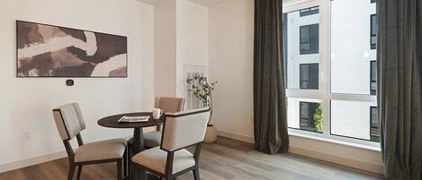 A modern dining area with a round table, two chairs, a wall-mounted abstract painting, and a large window with curtains.
