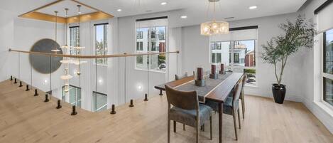 A modern dining area with a long wooden table, chairs, pendant lights, and a potted plant.