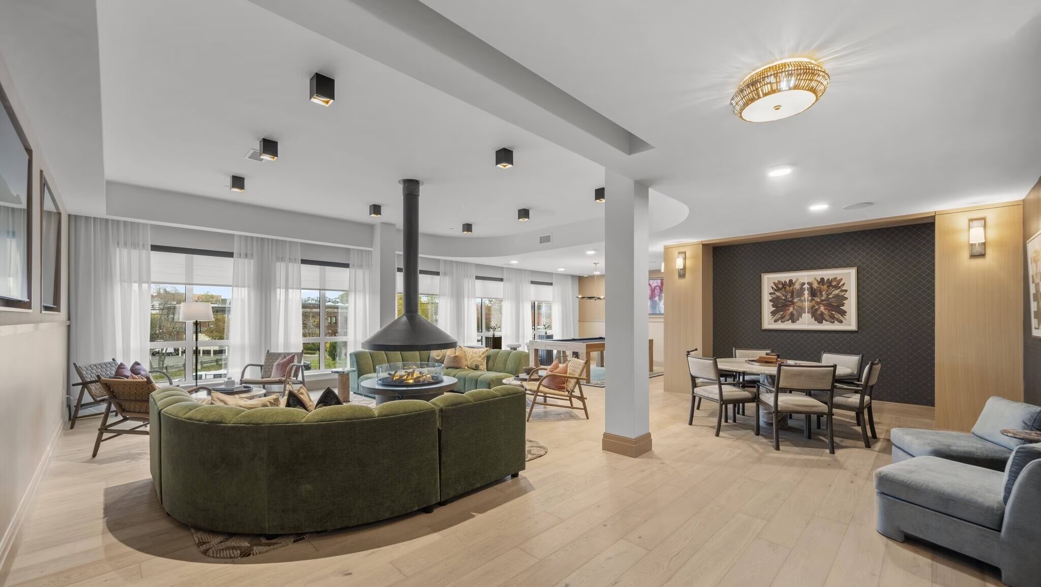 Een ruime woonkamer met een grote groene bank, een open haard, een eettafel met stoelen, en een groot raam met uitzicht.