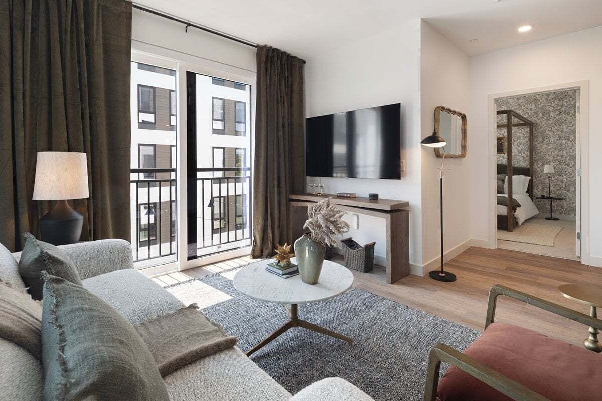 A modern living room with a sofa, a round coffee table, a flat-screen TV, and a balcony with curtains.