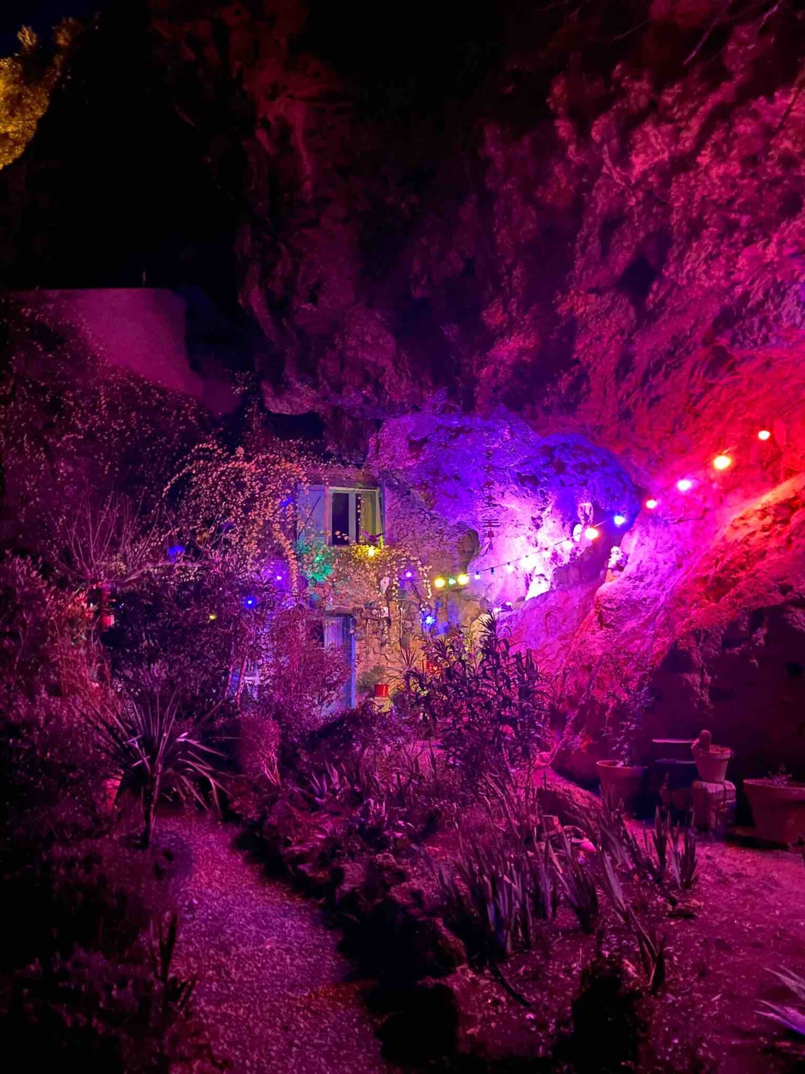 Eine höhlenartige Struktur mit einem kleinen Gebäude, beleuchtet von bunten Lichtern, umgeben von Vegetation.
