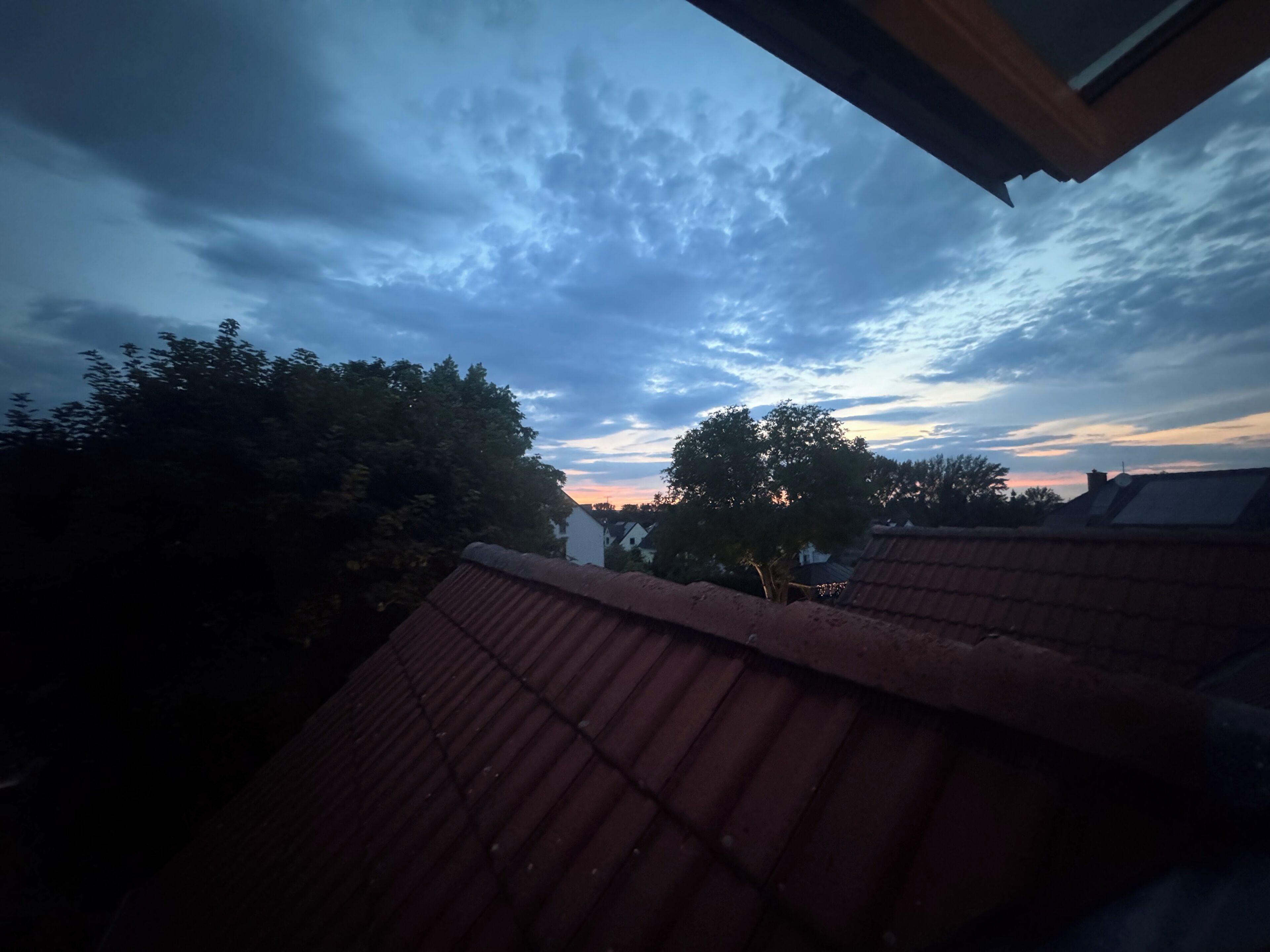 En vy över ett bostadsområde med ett hus som har ett rött tegeltak, träd och en molnig himmel.
