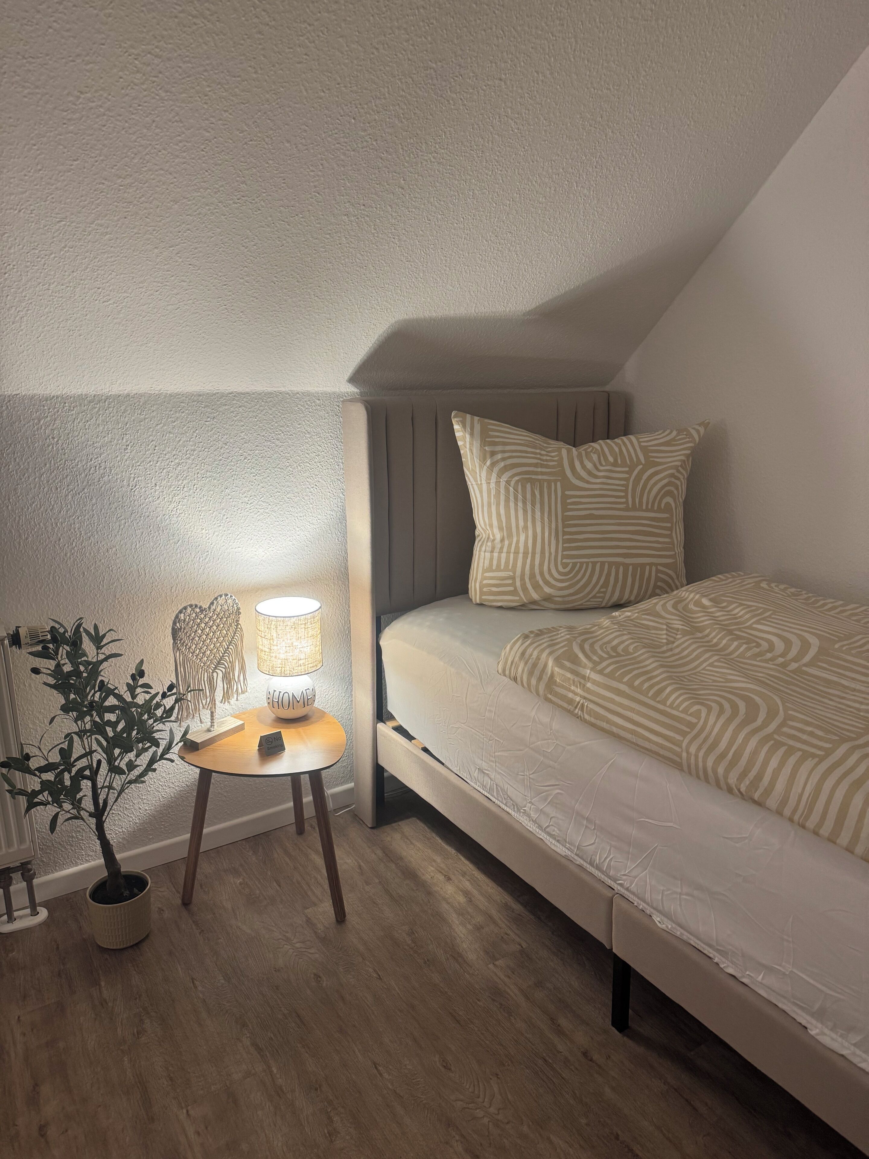 A bedroom with a sloped ceiling, a bed with striped bedding, a bedside table with a lamp, and a potted plant.