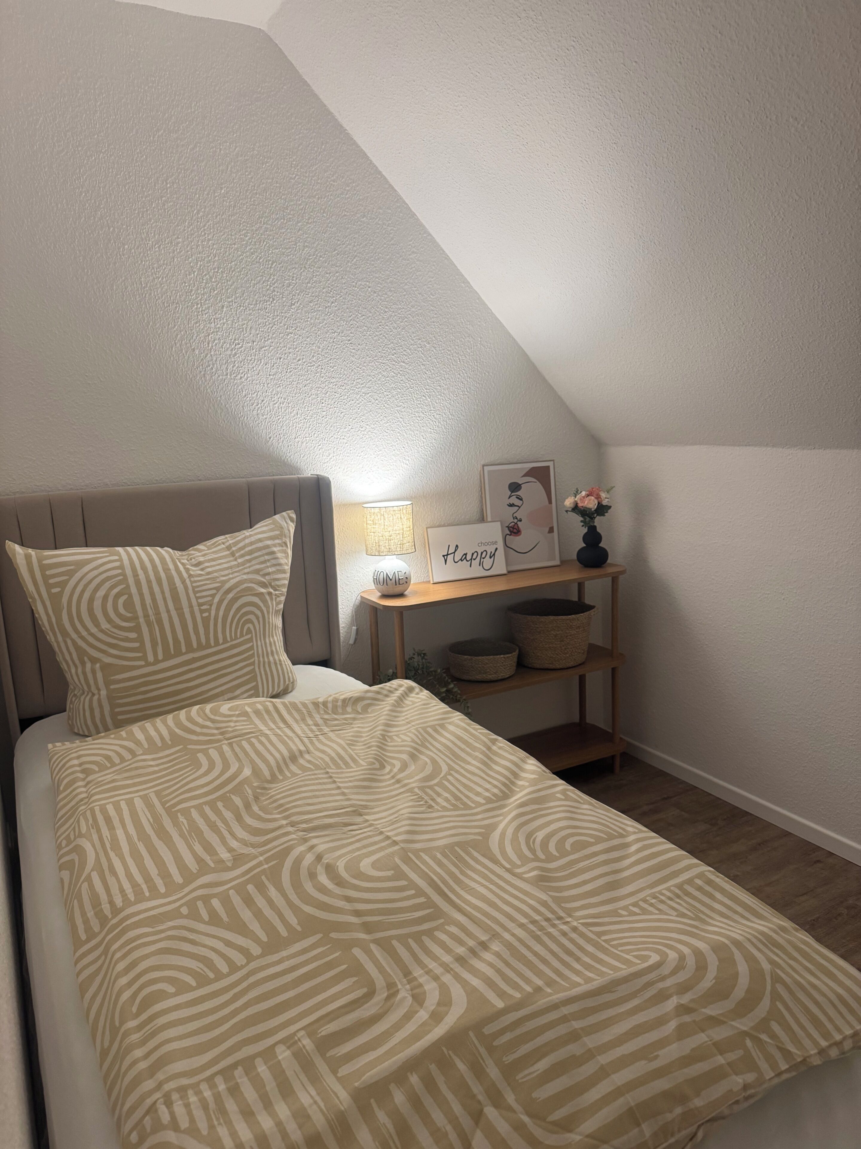 Ein Schlafzimmer mit einem Bett, Kissen, einem Nachttisch mit Lampe, einem gerahmten Bild und einer Vase mit Blumen.