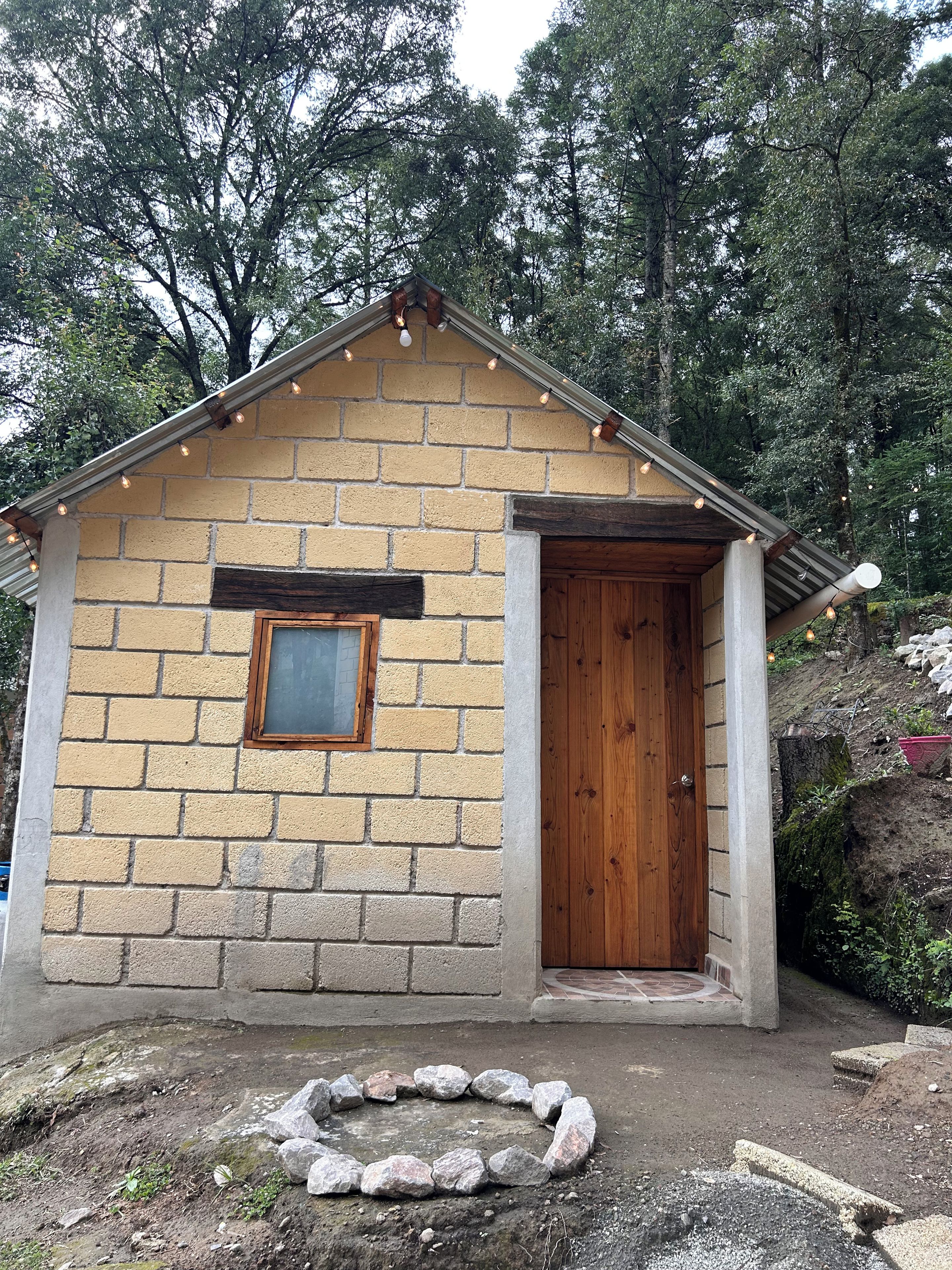 A small brick building with a wooden door and a window, surrounded by trees.