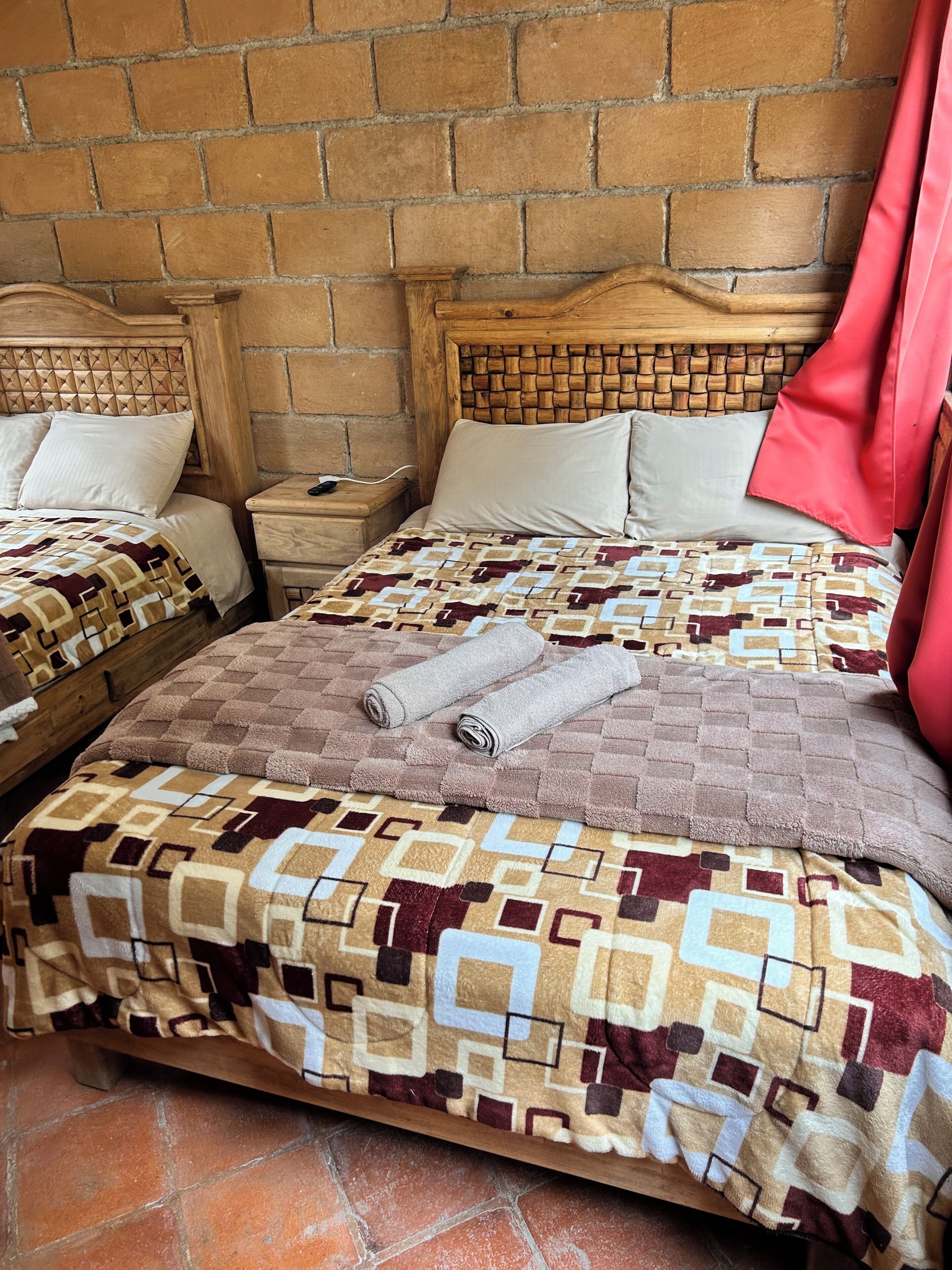 Quarto com parede de tijolos, cabeceira de madeira, colcha estampada e cortina vermelha.