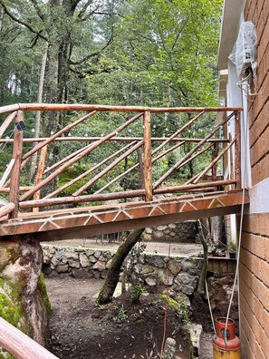 A rustic wooden bridge with a metal railing, leading to a stone wall and a building with a brick exterior.