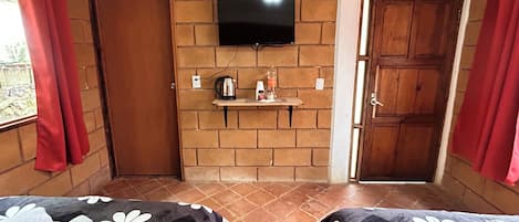 A room with two beds, a television mounted on a brick wall, a small shelf with items, and a door leading outside.