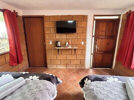 Una habitación con dos camas, un televisor instalado en una pared de ladrillo, un estante con objetos y una puerta que da al exterior.