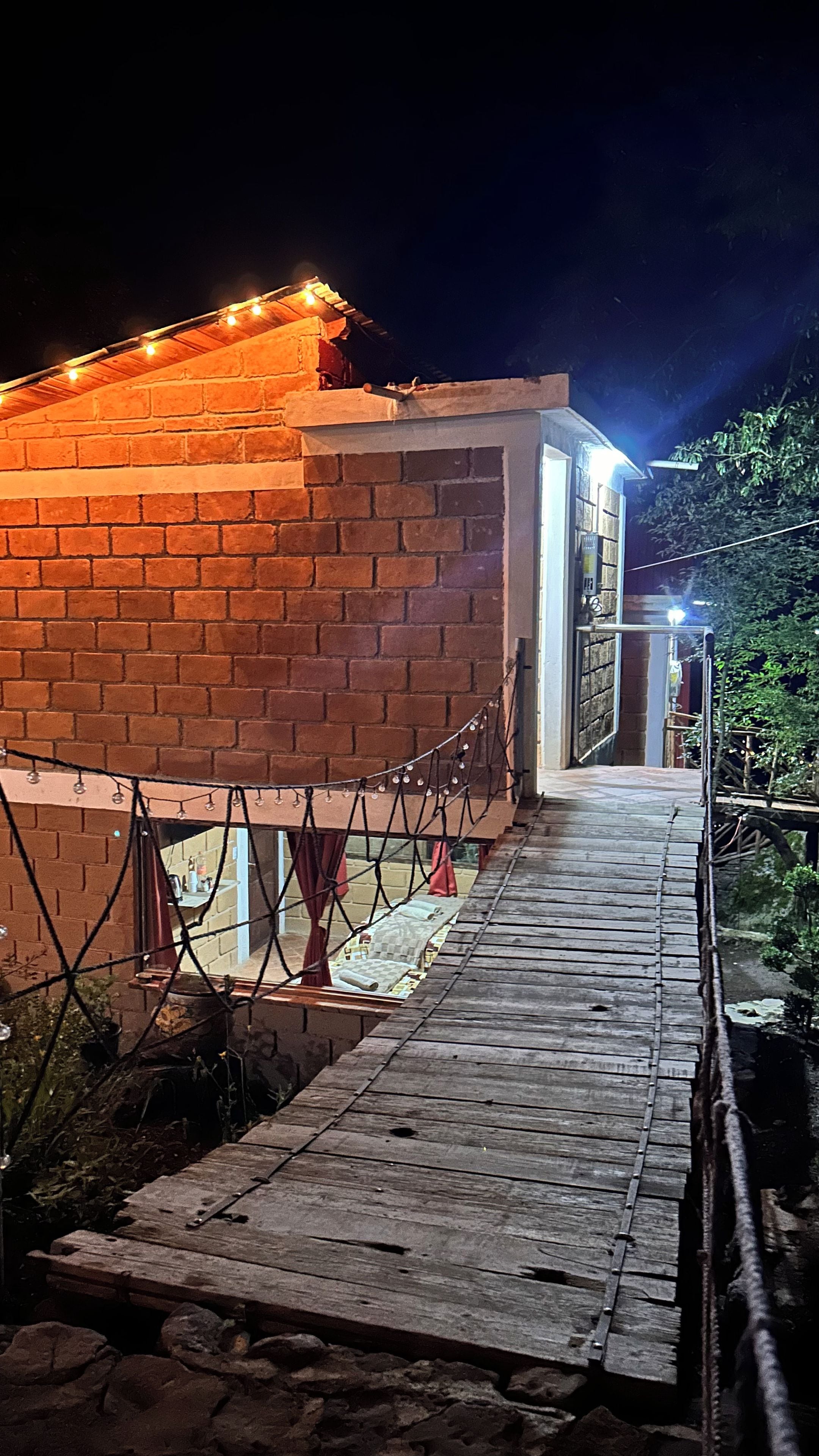 Un edificio de ladrillo con una terraza de madera que lleva a una puerta, rodeado de vegetación.