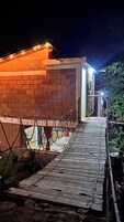 Edifício de tijolos com deck de madeira que leva a uma porta, rodeado por vegetação.
