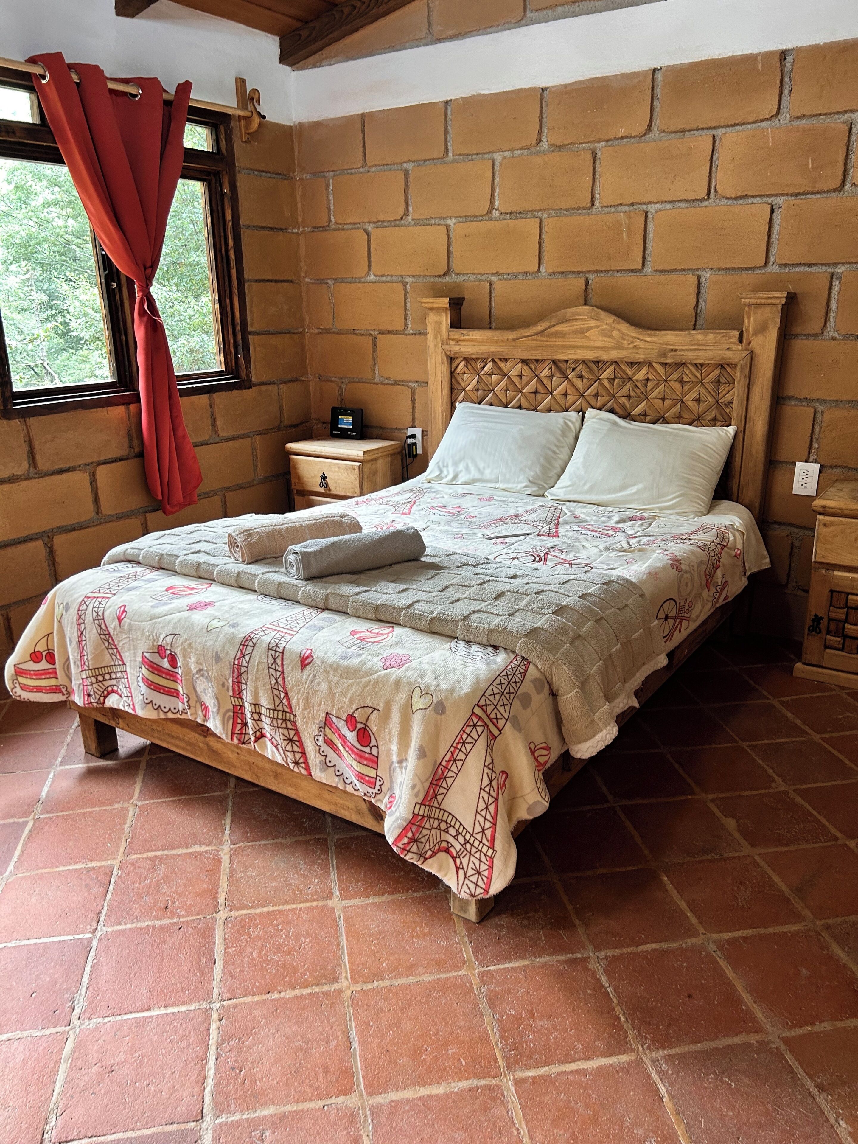 Una cama con una colcha estampada, un cabecero de madera, una mesita de noche con toma de corriente y una ventana con cortina roja.