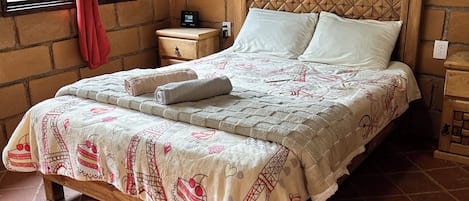 A bed with a patterned bedspread, a wooden headboard, a nightstand with a power outlet, and a window with a red curtain.