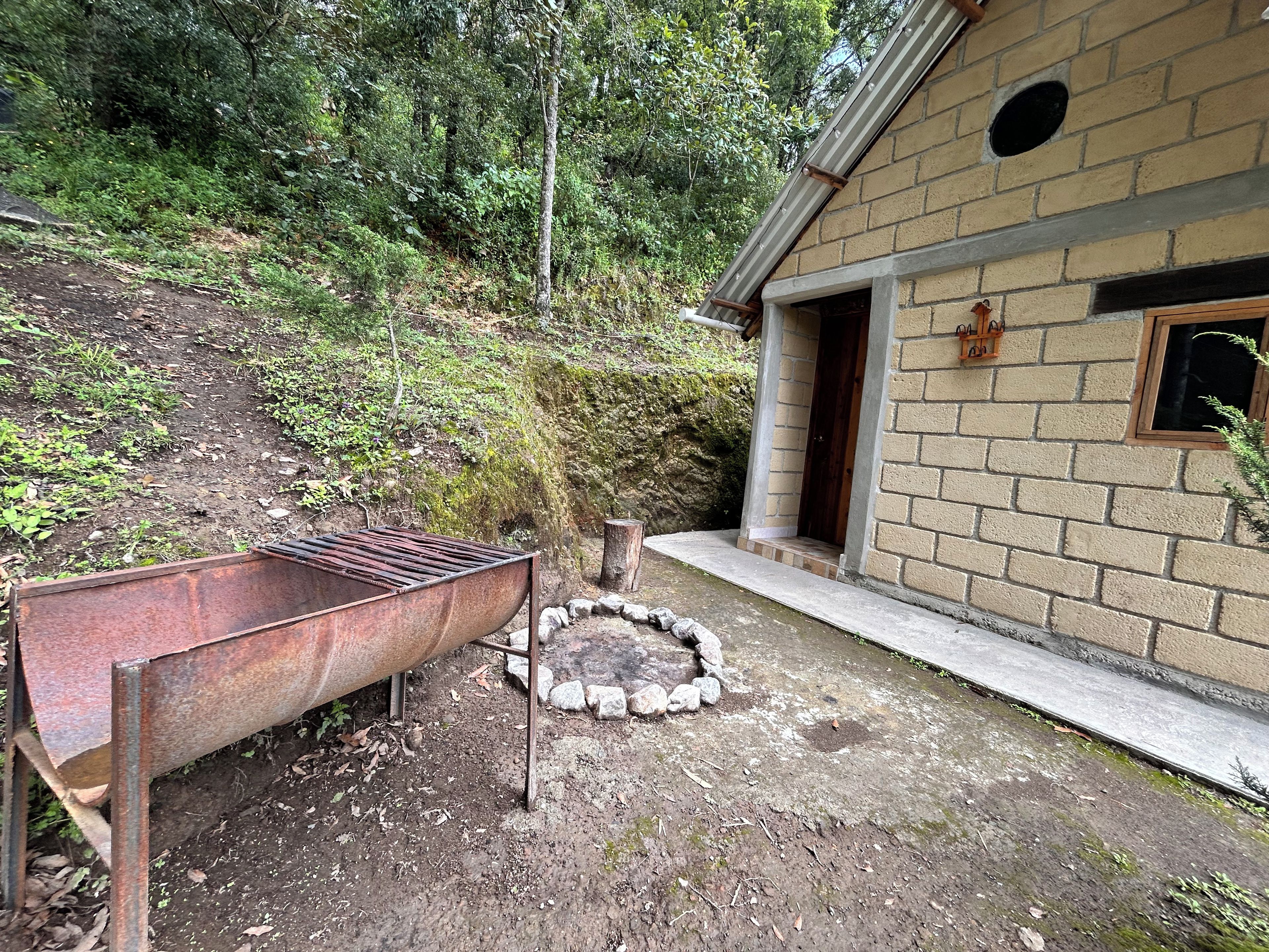 A rustic outdoor area with a metal barrel, a stone fire pit, and a brick building with a window.