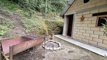 Uma área externa rústica com um barril de metal, um fosso de pedra e um edifício de tijolos com uma janela.