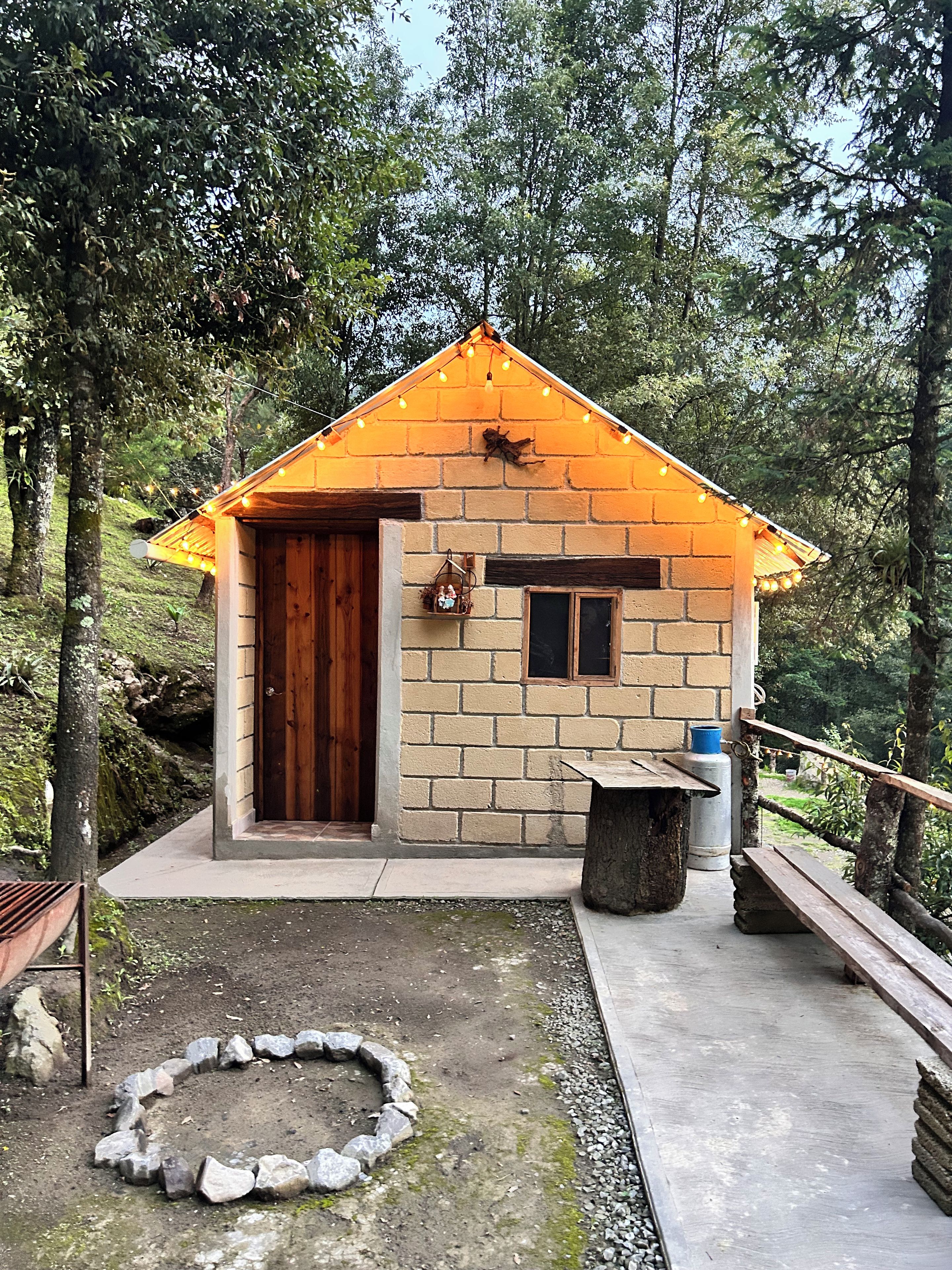 Una cabaña pequeña de una sola planta con paredes de ladrillo, una puerta de madera, un ventanal pequeño y un banco en el exterior.
