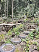 Zona boscosa con un puente de madera, muros de piedra y macetas de neumáticos.