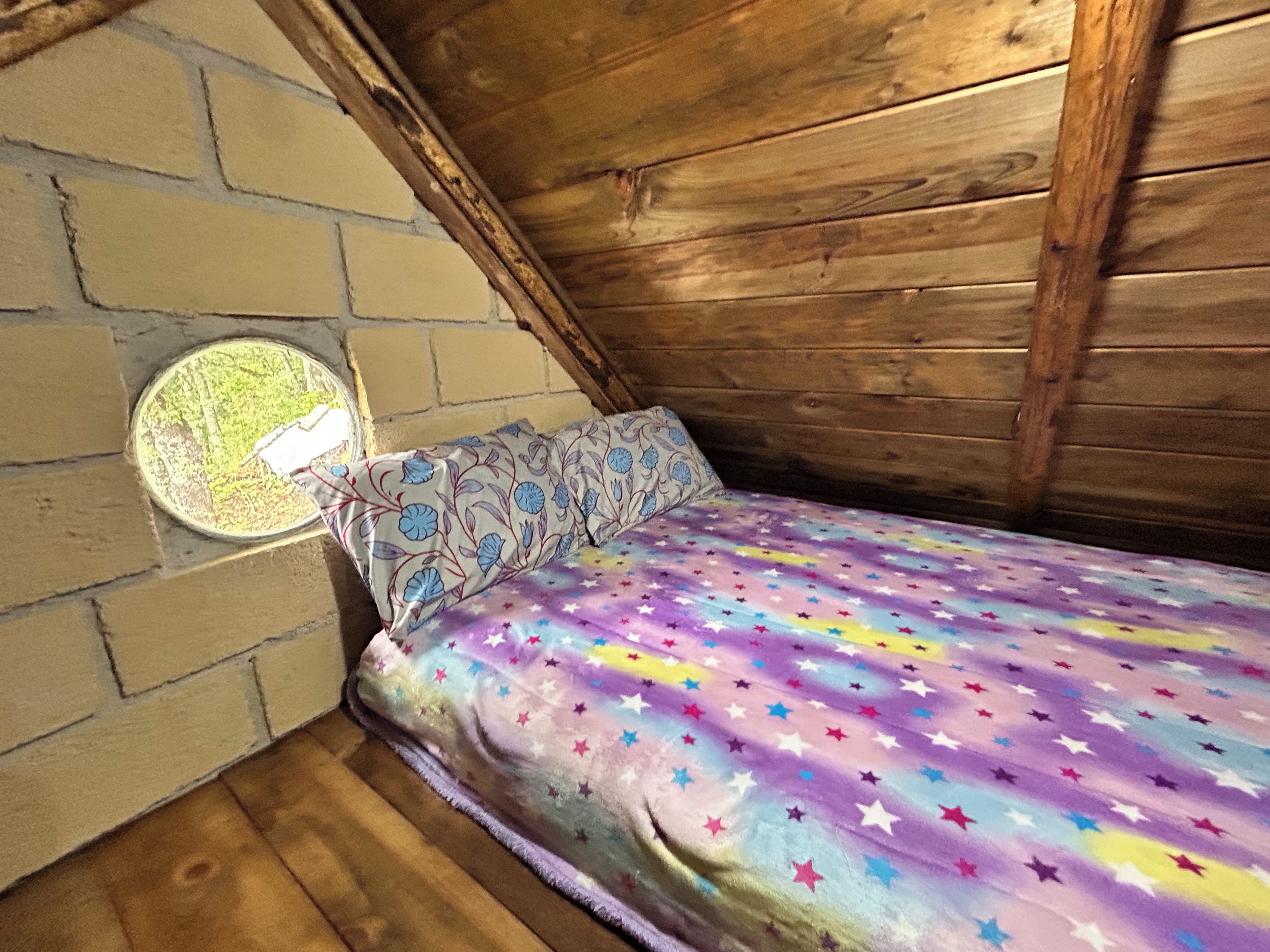 A small, cozy room with a sloped ceiling, wooden walls, a single bed with a colorful star-patterned bedspread, and a small window with a decorative frame.